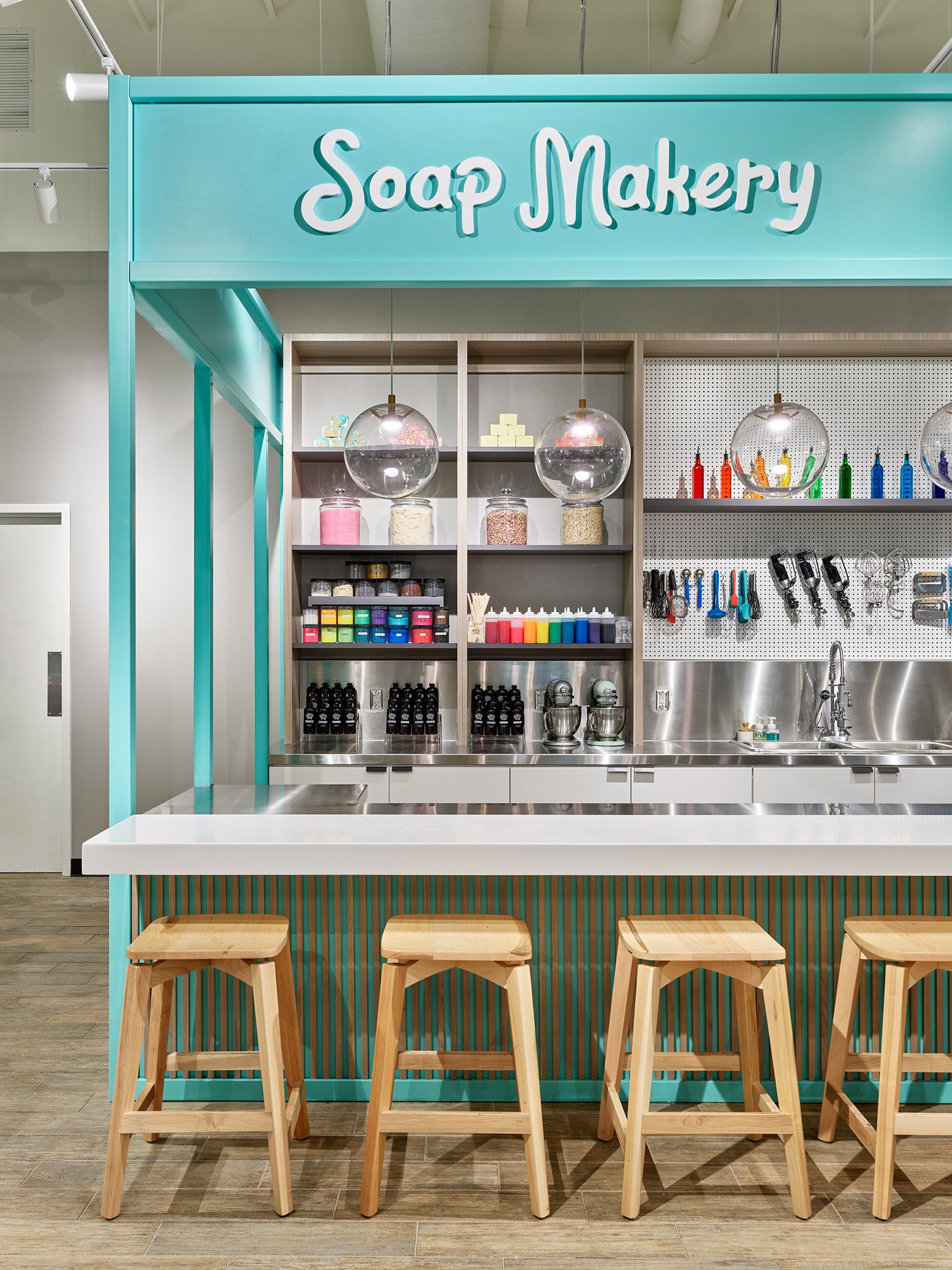 Buff City Soap teal Soap Makery counter with wooden stools on hardwood flooring.
