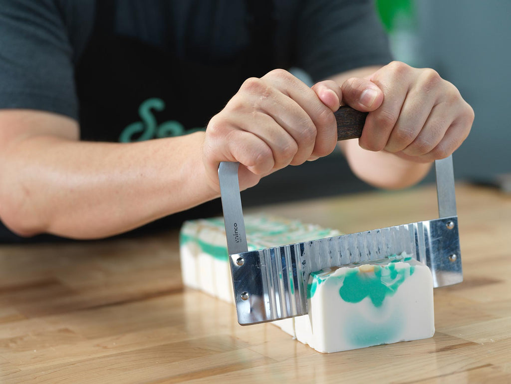 Buff City Soap maker cutting bars of soap on wooden table with metal tool.