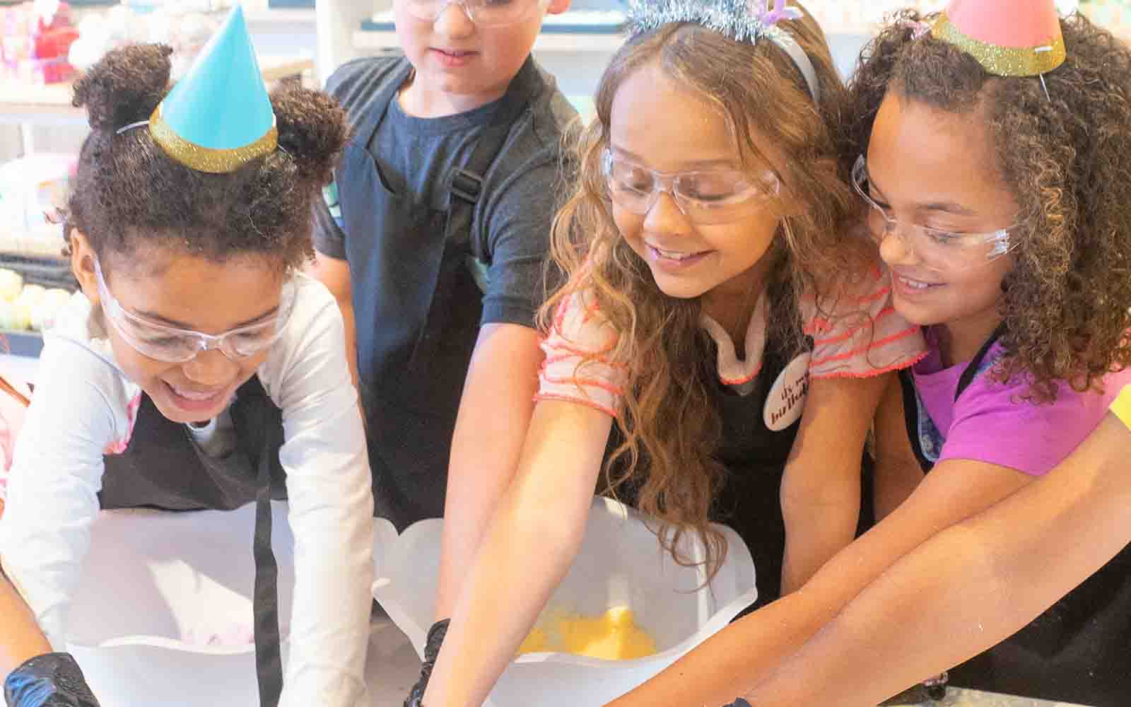 Four children making bath bombs at Buff City Soap while wearing protective glasses and party hats.