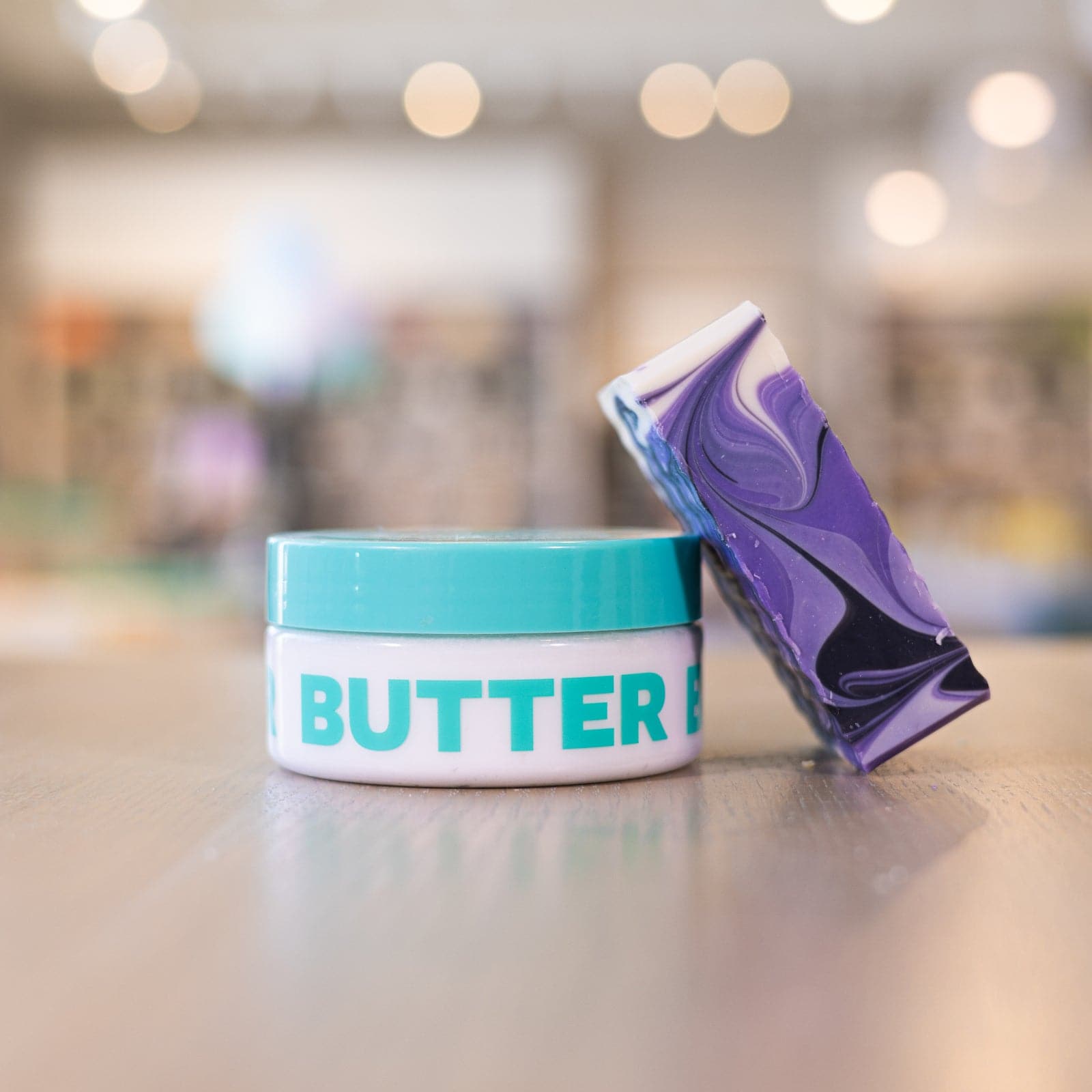 Purple and black soap bar leaning against clear body butter container with teal lid on counter